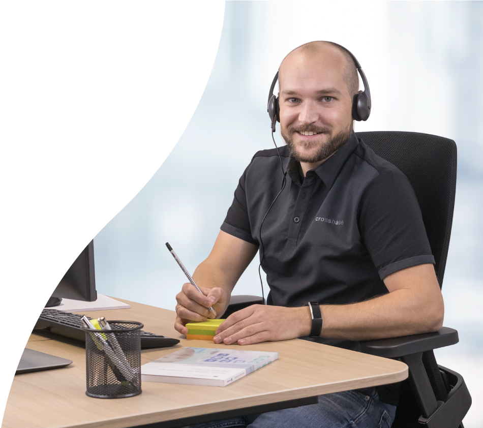 Ein crossbase-Mitarbeiter aus der Beratung blickt lächelnd in die Kamera. Er sitzt am Schreibtisch, ein Headset auf dem Kopf und mit Stift und Notizzettel-Block in der Hand.
