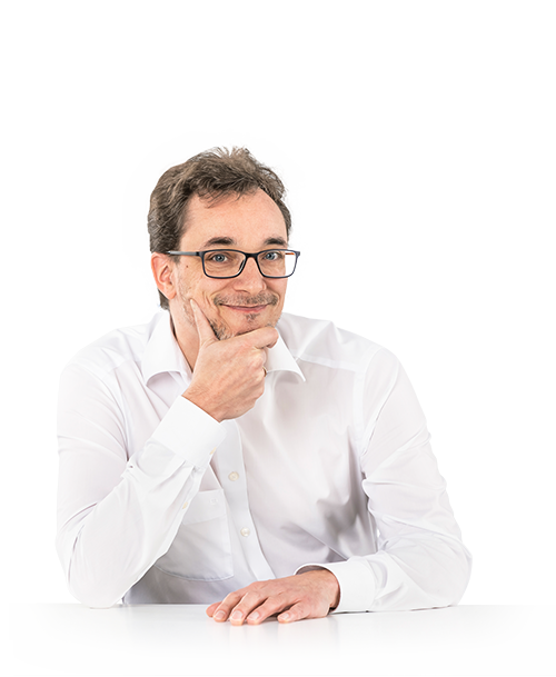 Jean-René Thies steht an einem Tisch, das Kinn auf eine Hand gestützt. Er trägt eine Brille und ein weißes Hemd und blickt lächelnd in die Kamera.