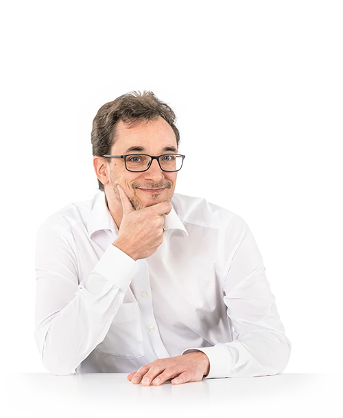 Jean-René Thies steht an einem Tisch, das Kinn auf eine Hand gestützt. Er trägt eine Brille und ein weißes Hemd und blickt lächelnd in die Kamera.