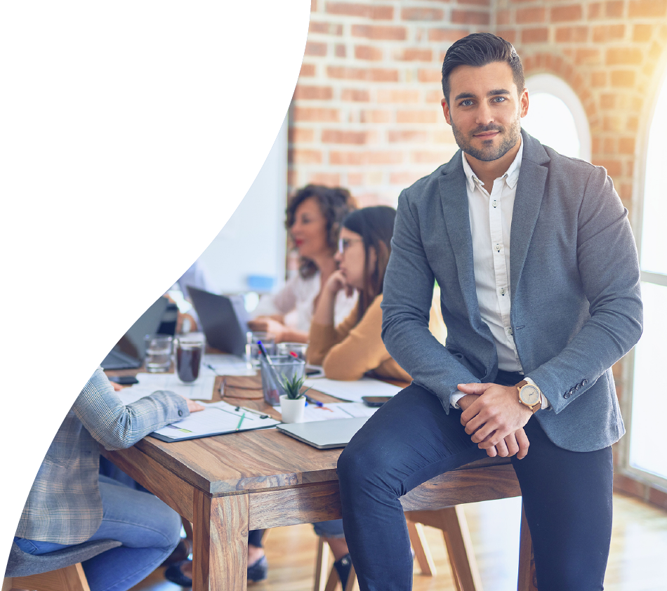 Ein junger Mann in Business-Outfit sitzt am Rand eines Konferenztisches und blickt direkt in die Kamera. Im Hintergrund arbeiten mehrere Personen konzentriert zusammen. Die Szene spielt in einem modernen Büro mit Ziegelwand.