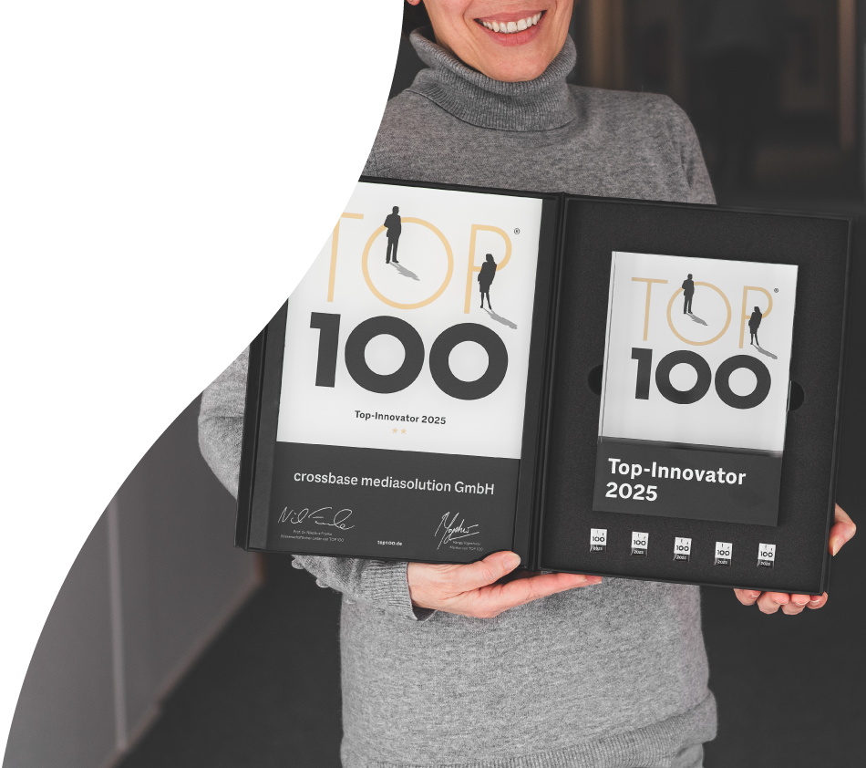 Eine Frau mit grauen Oberteil hält die unterzeichnete TOP 100-Auszeichnung für crossbase in den Händen und zeigt sie in die Kamera. Ihr Kopf ist nicht ganz zu sehen aber sie lächelt breit.