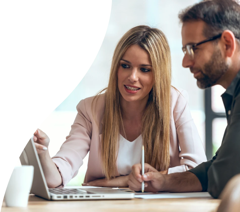 Two people, a woman and a man, sit together at a table and look intently at a laptop screen. The woman points at the screen and explains something while the man takes notes. Both appear engrossed in their work or a presentation.