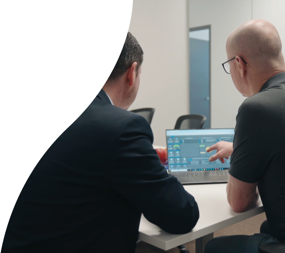 Two men are sitting with their backs to the camera in an office meeting room. On the table in front of them is a laptop with the crossbase dashboard open. The man on the right is pointing at the display and wearing glasses.