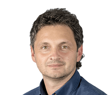 Reinhard Sutterlüty from meusburger smiles at the camera. He has curly, dark hair and is wearing a dark blue shirt. The background shows a modern wave shape in white, light blue and dark blue, which curves from the bottom left to the top right of the picture.