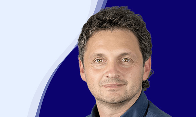 Reinhard Sutterlüty from meusburger smiles at the camera. He has curly, dark hair and is wearing a dark blue shirt. The background shows a modern wave shape in white, light blue and dark blue, which curves from the bottom left to the top right of the picture.
