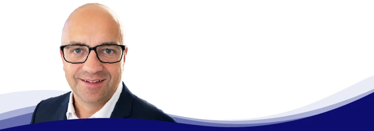 Tom Ruegenberg, Project Manager at Gebr. Kemper, smiles and looks into the camera. He wears black-rimmed glasses, a light blue shirt with an open collar and a gray jacket over it. He is bald and clean-shaven.