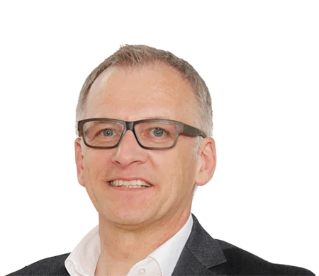 A middle-aged man looks into the camera with a smile. He has short, gray hair and wears dark glasses with distinctive rectangular frames. He is dressed in a white shirt with an open collar and a dark jacket.