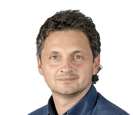 Reinhard Sutterlüty from meusburger smiles at the camera. He has curly, dark hair and is wearing a dark blue shirt. The background shows a modern wave shape in white, light blue and dark blue, which curves from the bottom left to the top right of the picture.