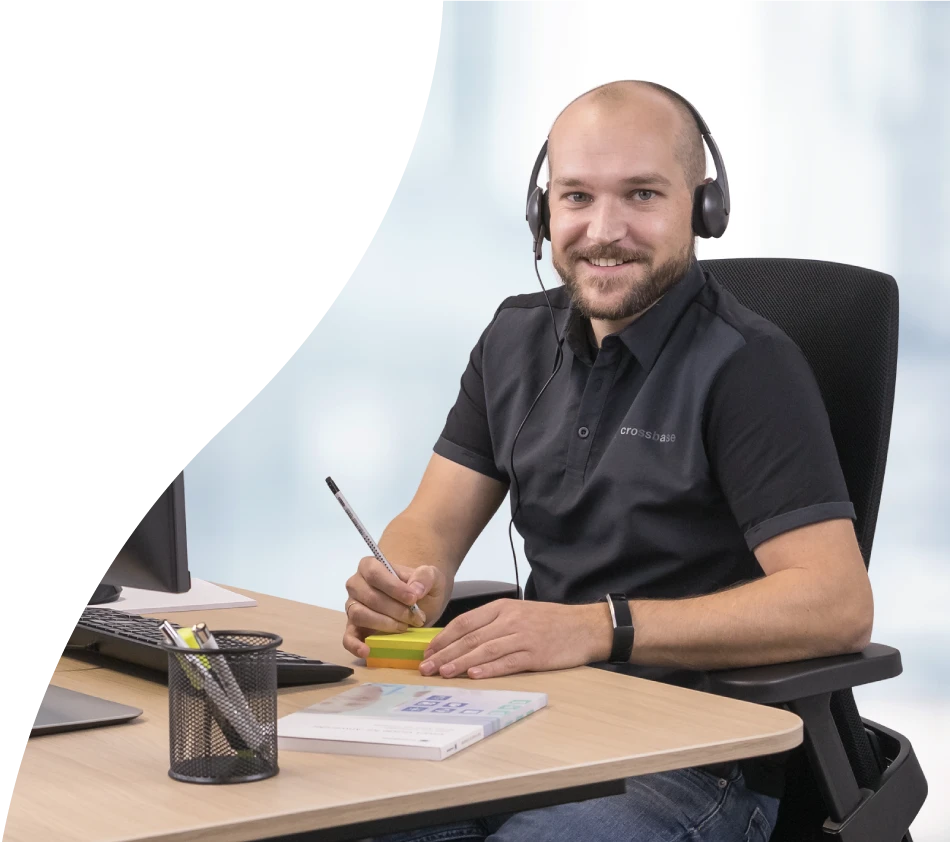 A crossbase employee from the consulting department looks into the camera with a smile. He is sitting at his desk, a headset on his head and a pen and notepad in his hand.