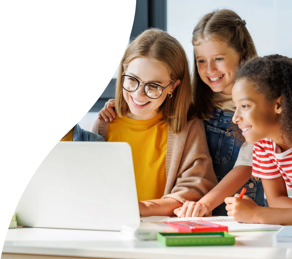 A woman with a pageboy cut and glasses looks at the screen of a laptop with a smile. Surrounding her are boys and girls of around six or seven years old, who are looking at the monitor with interest and happiness.