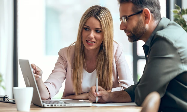 Two people, a woman and a man, sit together at a table and look intently at a laptop screen. The woman points at the screen and explains something while the man takes notes. Both appear engrossed in their work or a presentation.