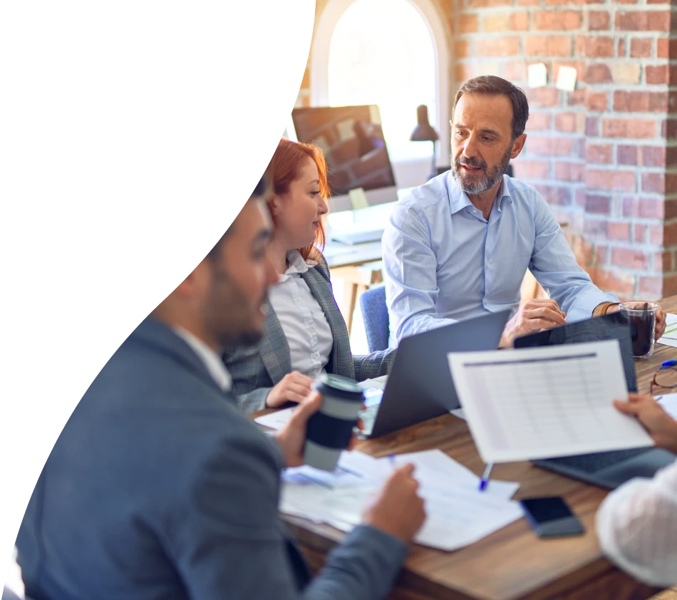 Quatre personnes en tenue d'affaires sont assises à une table en bois dans un bureau moderne. Ils sont plongés dans une conversation et travaillent ensemble avec des ordinateurs portables, des documents et des tasses de café. En arrière-plan, on aperçoit une fenêtre avec la lumière du jour ainsi qu'un écran. L'atmosphère est concentrée et professionnelle.