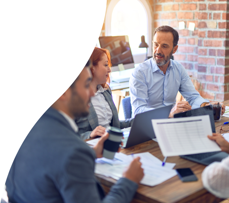 Quatre personnes en tenue d'affaires sont assises à une table en bois dans un bureau moderne. Ils sont plongés dans une conversation et travaillent ensemble avec des ordinateurs portables, des documents et des tasses de café. En arrière-plan, on aperçoit une fenêtre avec la lumière du jour ainsi qu'un écran. L'atmosphère est concentrée et professionnelle.