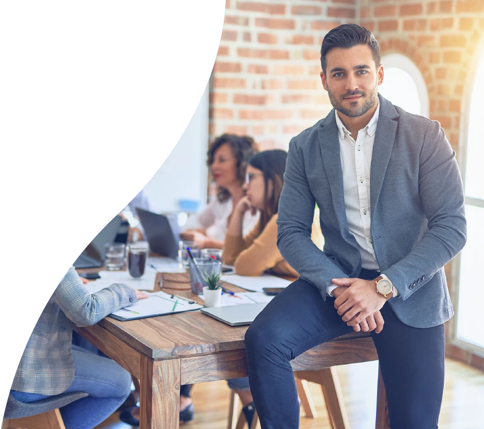 Un jeune homme en tenue d'affaires est assis au bord d'une table de conférence et regarde directement la caméra. En arrière-plan, plusieurs personnes travaillent ensemble de manière concentrée. La scène se déroule dans un bureau moderne avec un mur de briques.