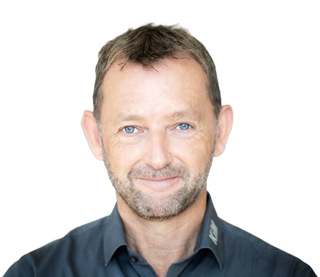 Achim Angel, directeur de KiM GmbH - Member of 4PACE, sourit aimablement à la caméra. Il a les cheveux bruns courts, une courte barbe et porte une chemise gris foncé avec un col légèrement ouvert. En arrière-plan, une forme ondulée incurvée en blanc, bleu clair et bleu foncé confère à la photo un design moderne.