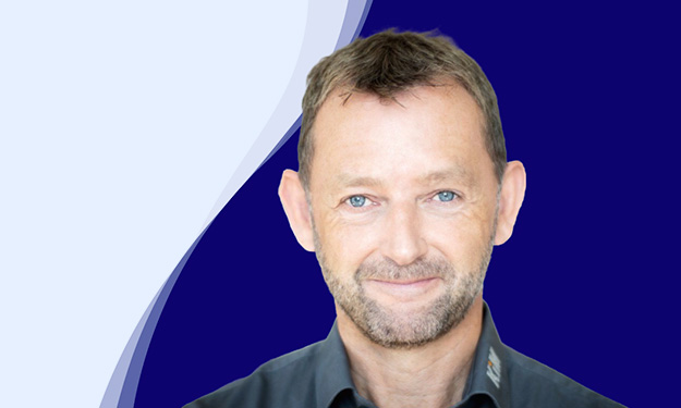 Achim Angel, directeur de KiM GmbH - Member of 4PACE, sourit aimablement à la caméra. Il a les cheveux bruns courts, une courte barbe et porte une chemise gris foncé avec un col légèrement ouvert. En arrière-plan, une forme ondulée incurvée en blanc, bleu clair et bleu foncé confère à la photo un design moderne.