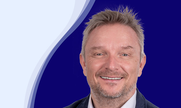 Holger Geck, directeur du marketing et de la gestion des données produits chez Gust. Alberts, sourit à la caméra. Il a les cheveux courts, blond grisâtre et une courte barbe. Il porte une chemise blanche et une veste sombre. En arrière-plan, on peut voir une forme de vague incurvée en blanc, bleu clair et bleu foncé.