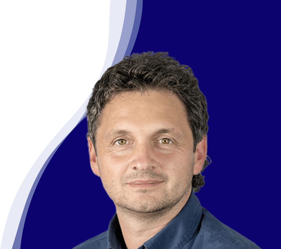 Reinhard Sutterlüty de l'entreprise meusburger sourit à la caméra. Il a les cheveux bouclés et foncés et porte une chemise bleu foncé. L'arrière-plan montre une forme de vague moderne en blanc, bleu clair et bleu foncé, qui s'étend en courbe du bas à gauche au haut à droite de l'image.